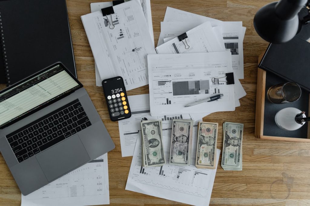 a-laptop-near-the-dollars-and-papers-on-a-wooden-table-6693655 Overhead view of financial documents, cash, and technology on a wooden desk.