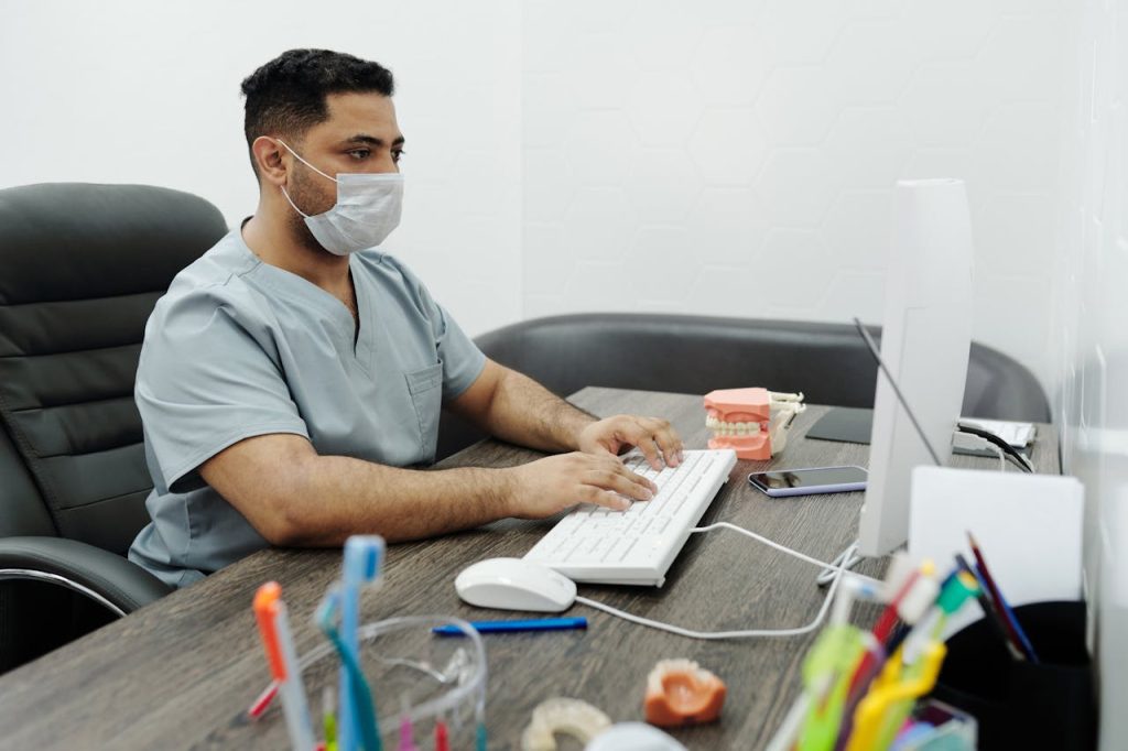 a-man-wearing-a-face-mask-using-computer-4269505 Dentist wearing mask working on computer at office desk with dental model.