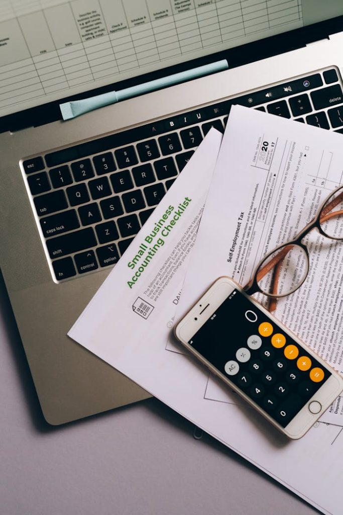 a-mobile-phone-and-documents-near-the-laptop-8962458 Small business accounting setup featuring a laptop, documents, and smartphone calculator.